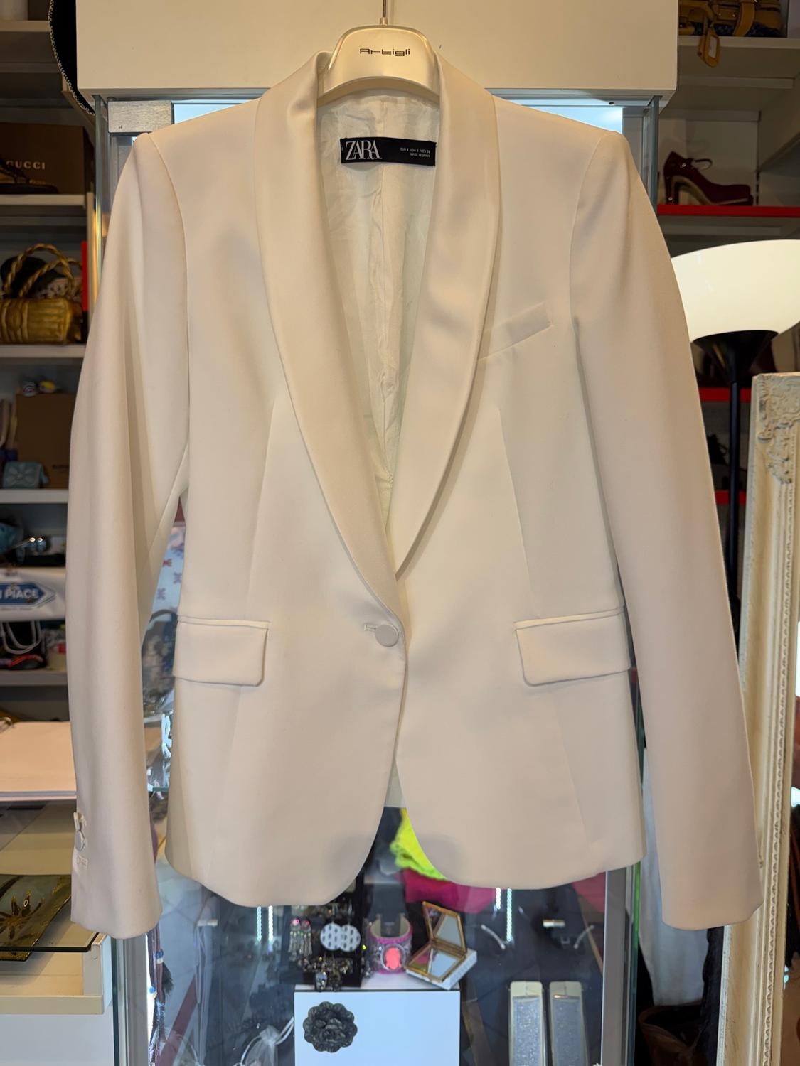 White Zara jacket hanging on a hanger, with a shop background showing accessories and shoes on shelves.