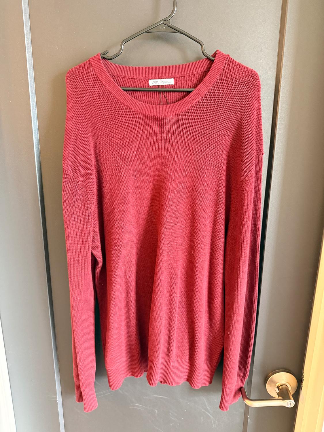 Red knit sweater hanging on a hanger over a gray door. Visible tag on the collar.