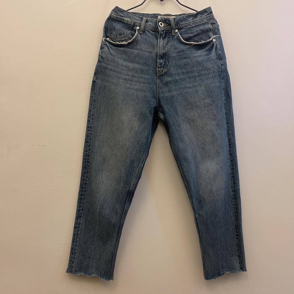 Worn blue jeans hanging on a hanger against a beige wall. They have front pockets and frayed hems.