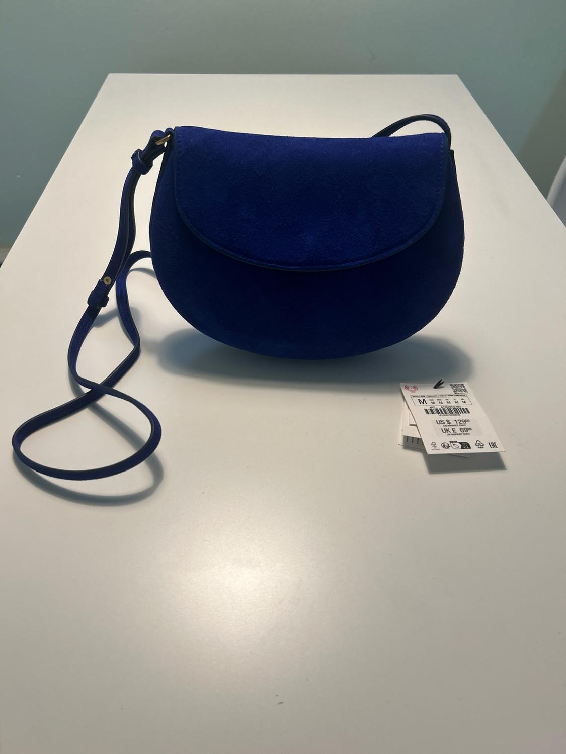 Dark blue flap shoulder bag on a white table. Label with text: M US $ 129.00 UK £ 69.00.