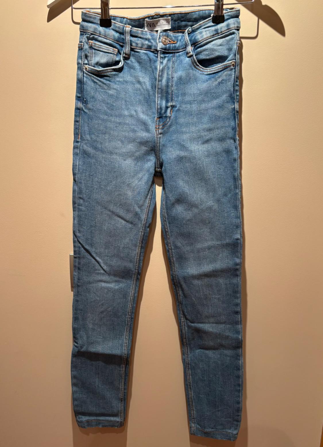 Light blue jeans hanging on a metal hanger against a beige background, with visible front and back pockets and a gold button.