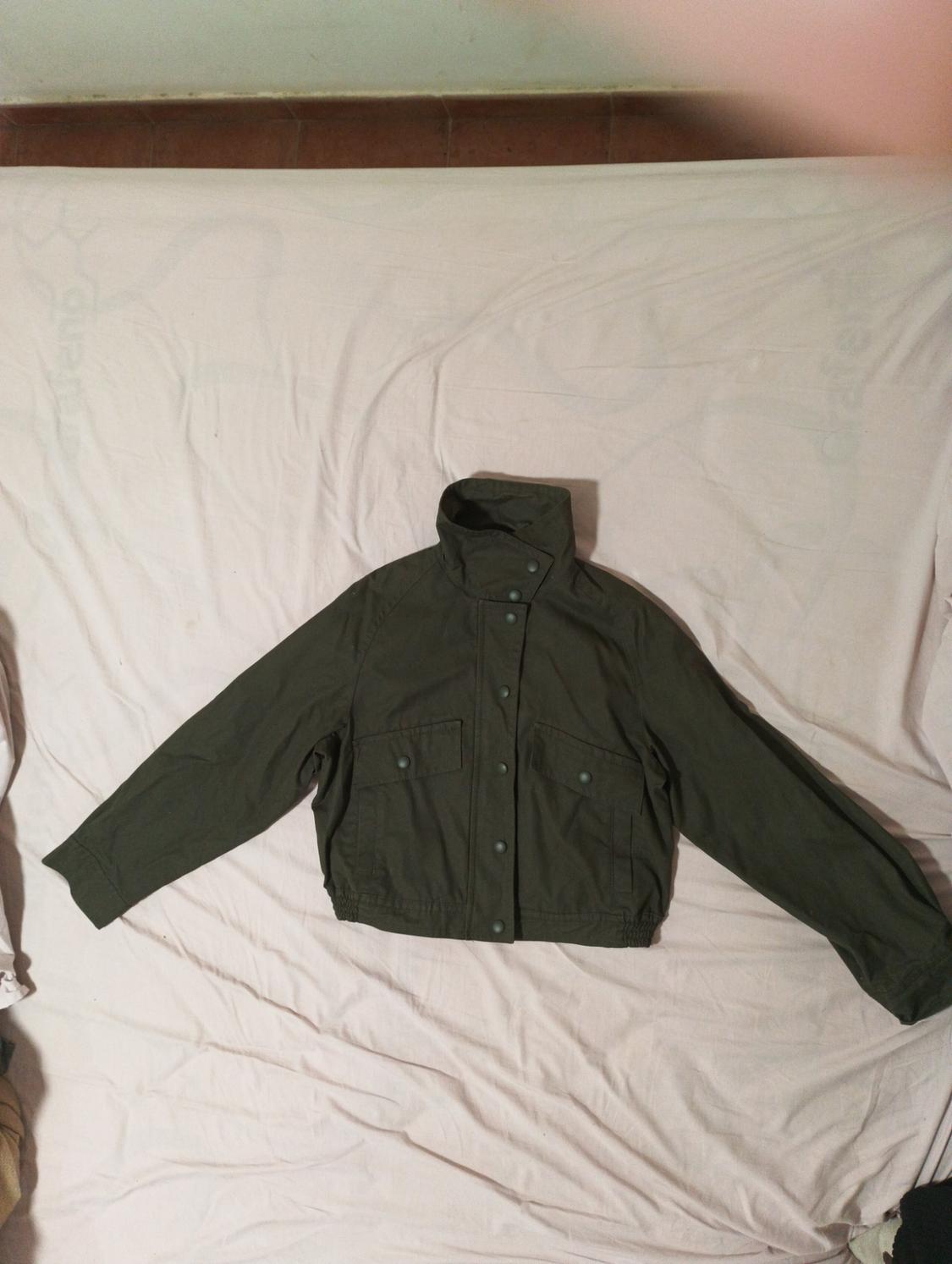 Olive green jacket with buttons and front pockets, placed on a bed with wrinkled white sheets.