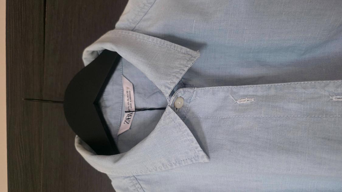 Light blue casual shirt in the foreground on a black hanger against a wooden background. Text: EUR XL USA XL MEX 42 MADE IN MOROCCO ZARA.