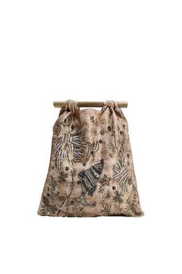 Beige handbag with floral and fish embroidery in metallic tones, with a wooden handle. White background.