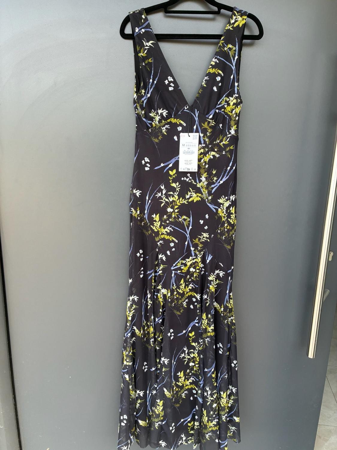 Long dark dress with a V-neckline and a floral print in shades of green, white and blue. It is hanging on a hanger against a grey background.