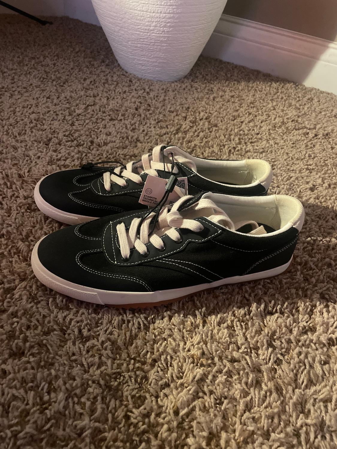Black trainers with white laces on a beige rug. Visible tag with "STANDARD MODEL" text. Background with white plant pot.