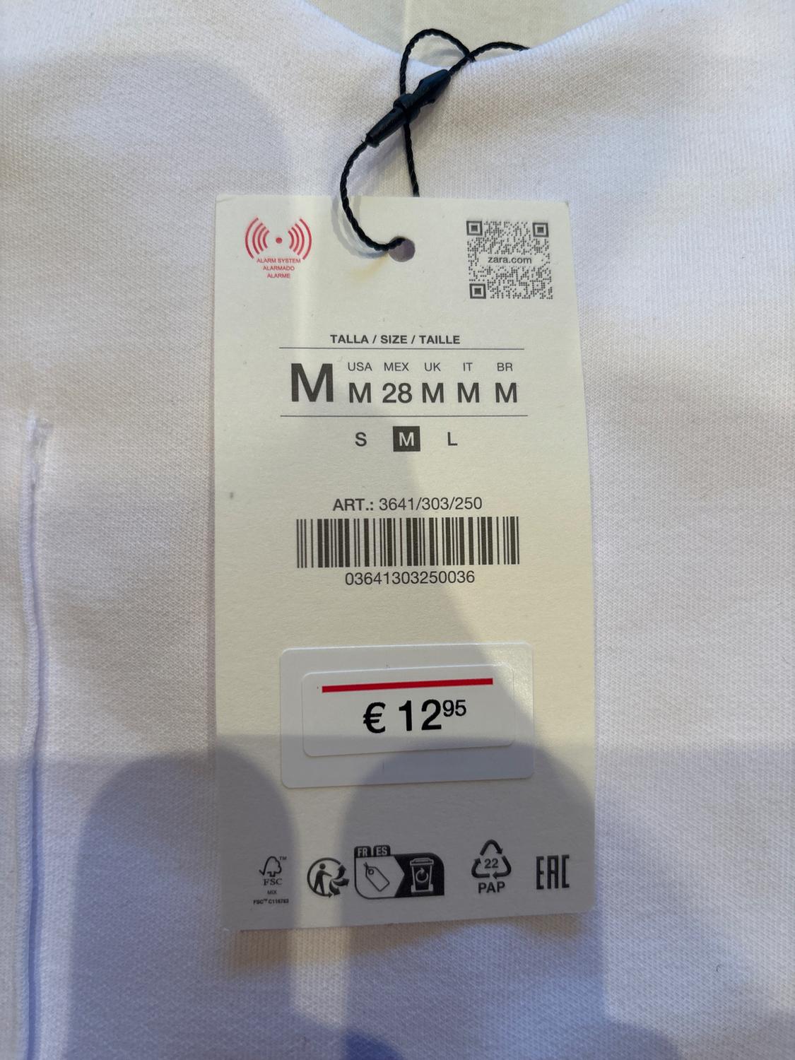 White garment tag with a price of €12.95, size M available in various nations, includes barcode and QR. White fabric background.