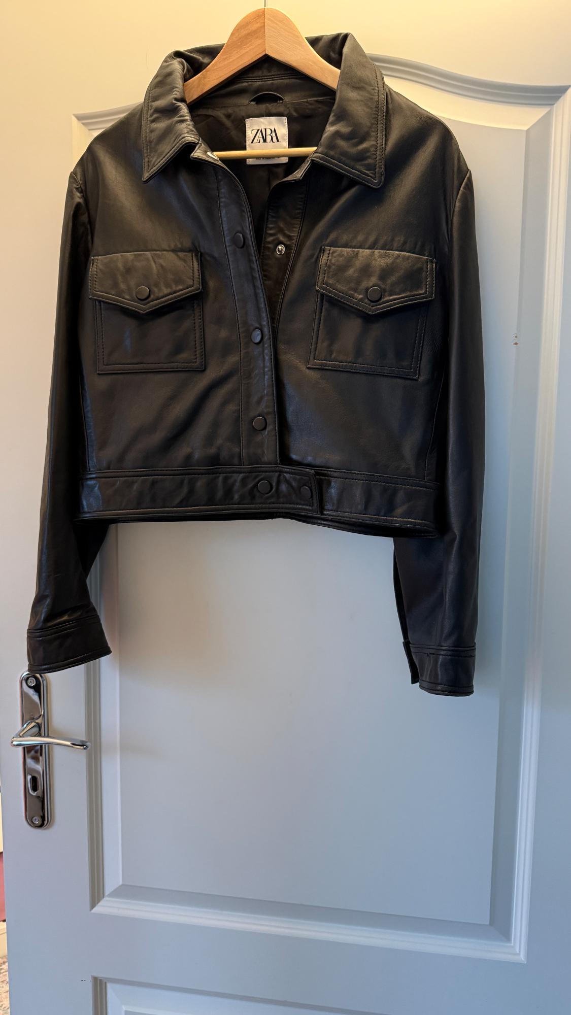 Black leather jacket hanging on a wooden hanger over a white door. Visible label with the text "ZARA".