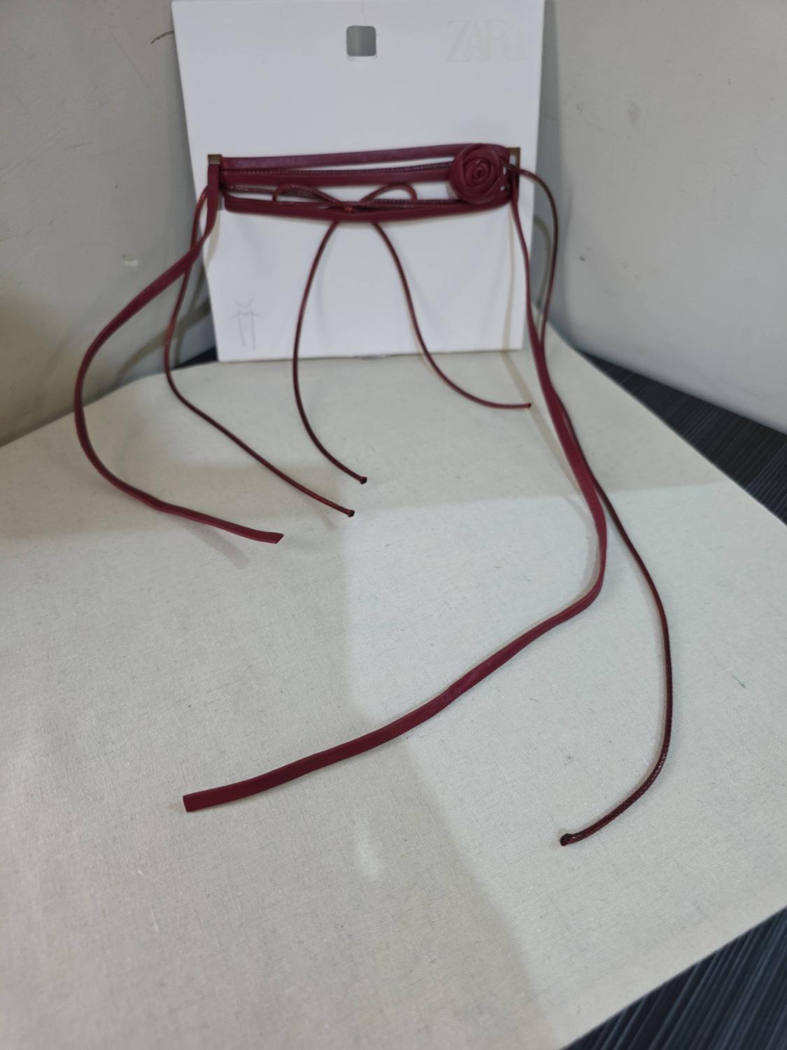 Burgundy strappy accessory with a flower detail on a white label bearing the text ZARA, on a light fabric surface.