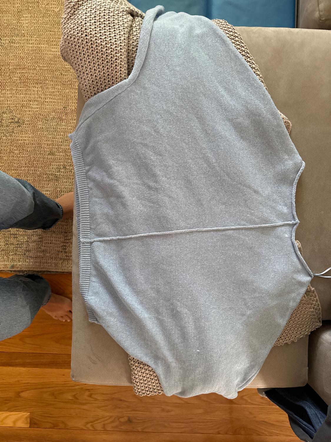 Light gray knit vest on a beige sofa, with a brown fabric underneath. Part of a leg visible in the lower left corner.