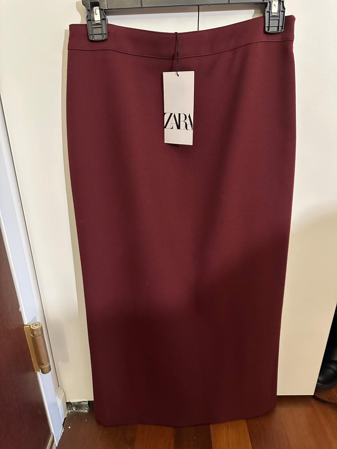 Burgundy long skirt hanging on a hanger with a ZARA tag, against a white wall background and wooden floor.