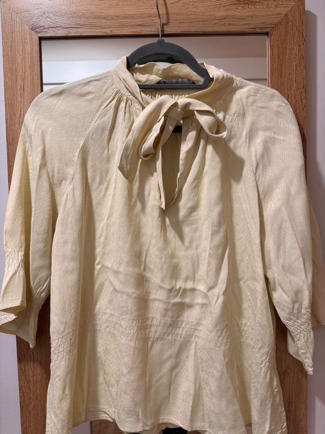 Beige three-quarter sleeve blouse with a bow at the neck, hanging on a black hanger in front of a wooden-framed mirror.