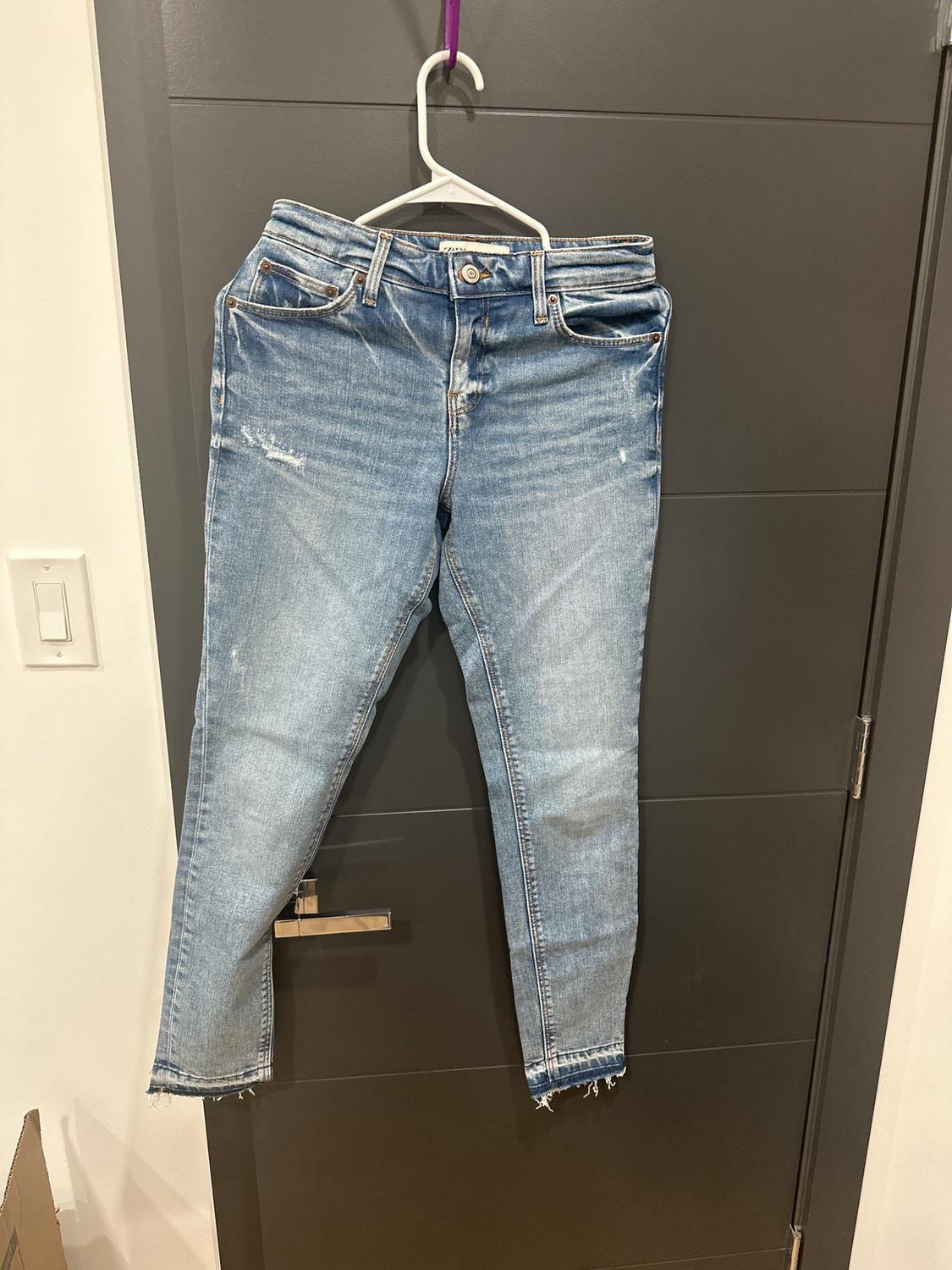 Light blue distressed jeans hanging on a white hanger over a gray door. Frayed details on the hems.