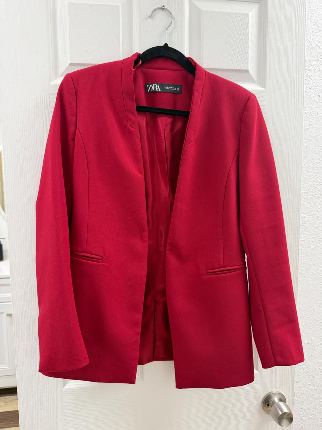 Red blazer with a ZARA tag hanging on a black hanger over a white door. Open-front, long-sleeved design with front pockets.