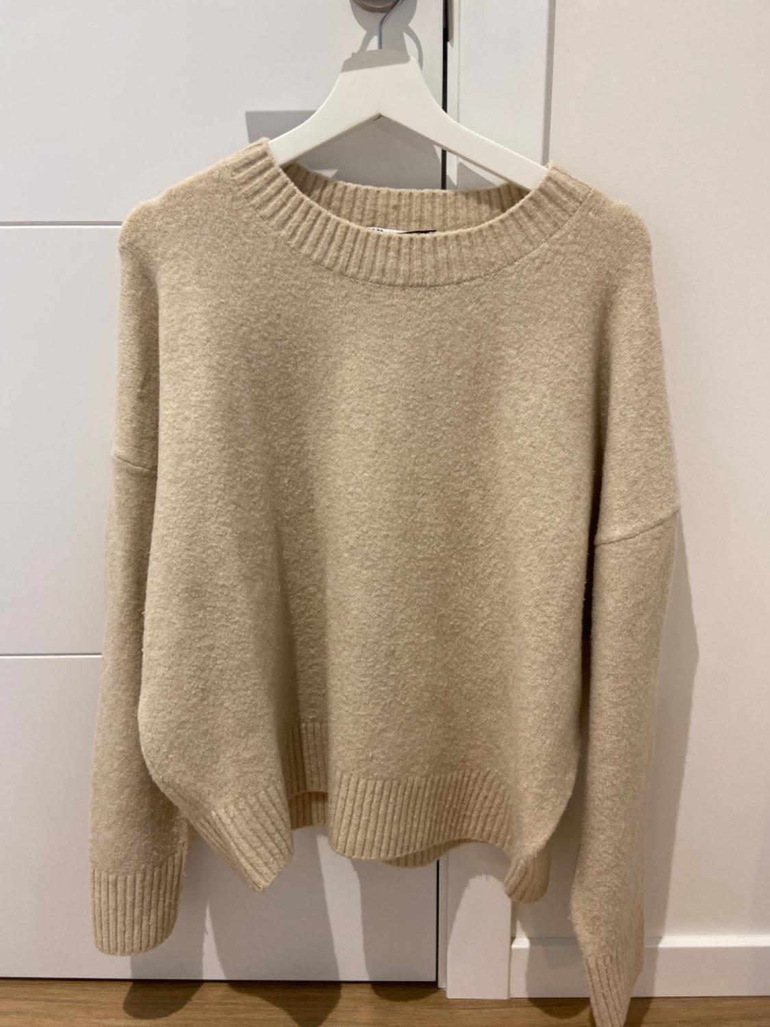 Beige chunky knit jumper hanging on a white hanger over a white door. Simple and cosy design.