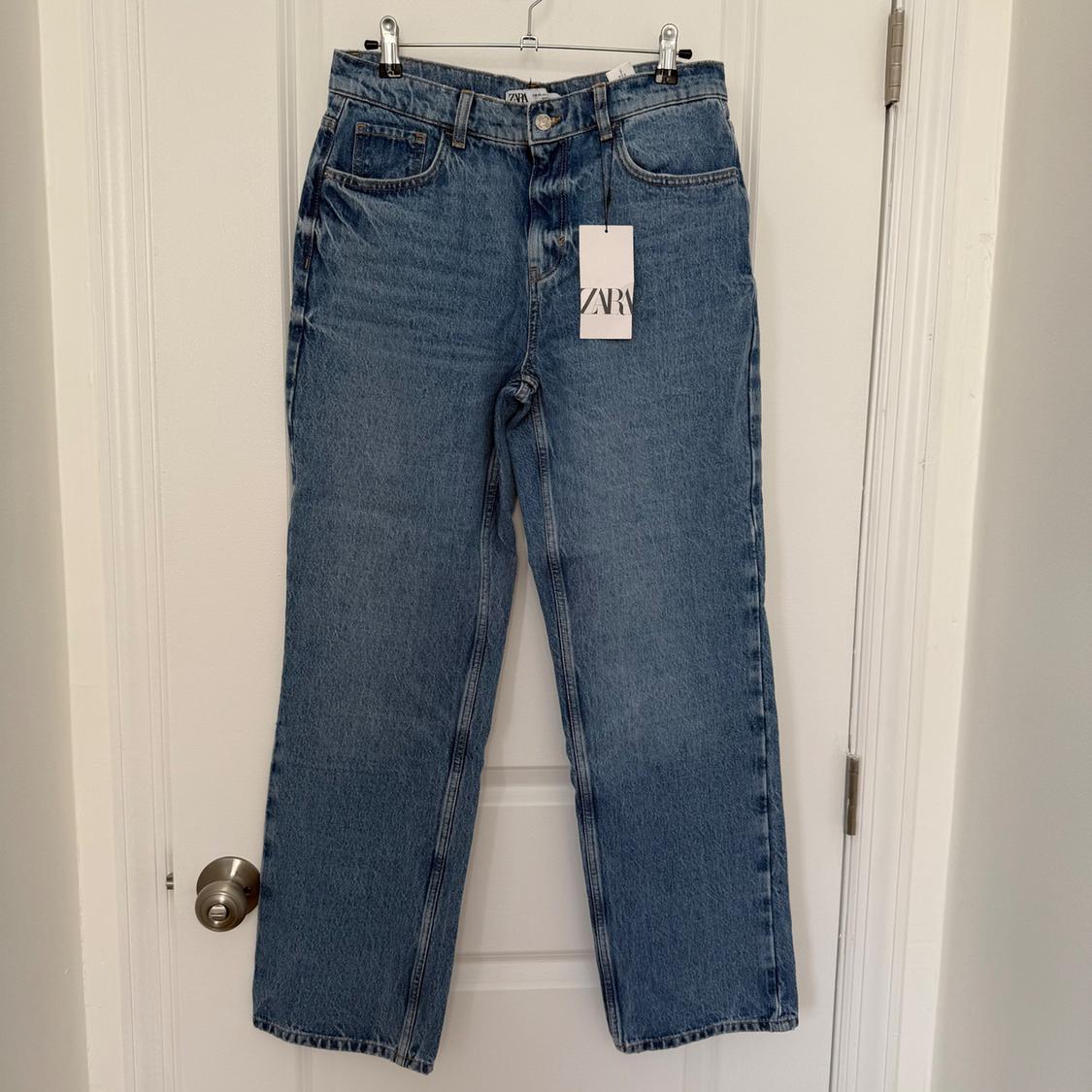 Light blue jeans hanging on a white door with a visible "ZARA" tag, minimalist background with a metal doorknob on the left.