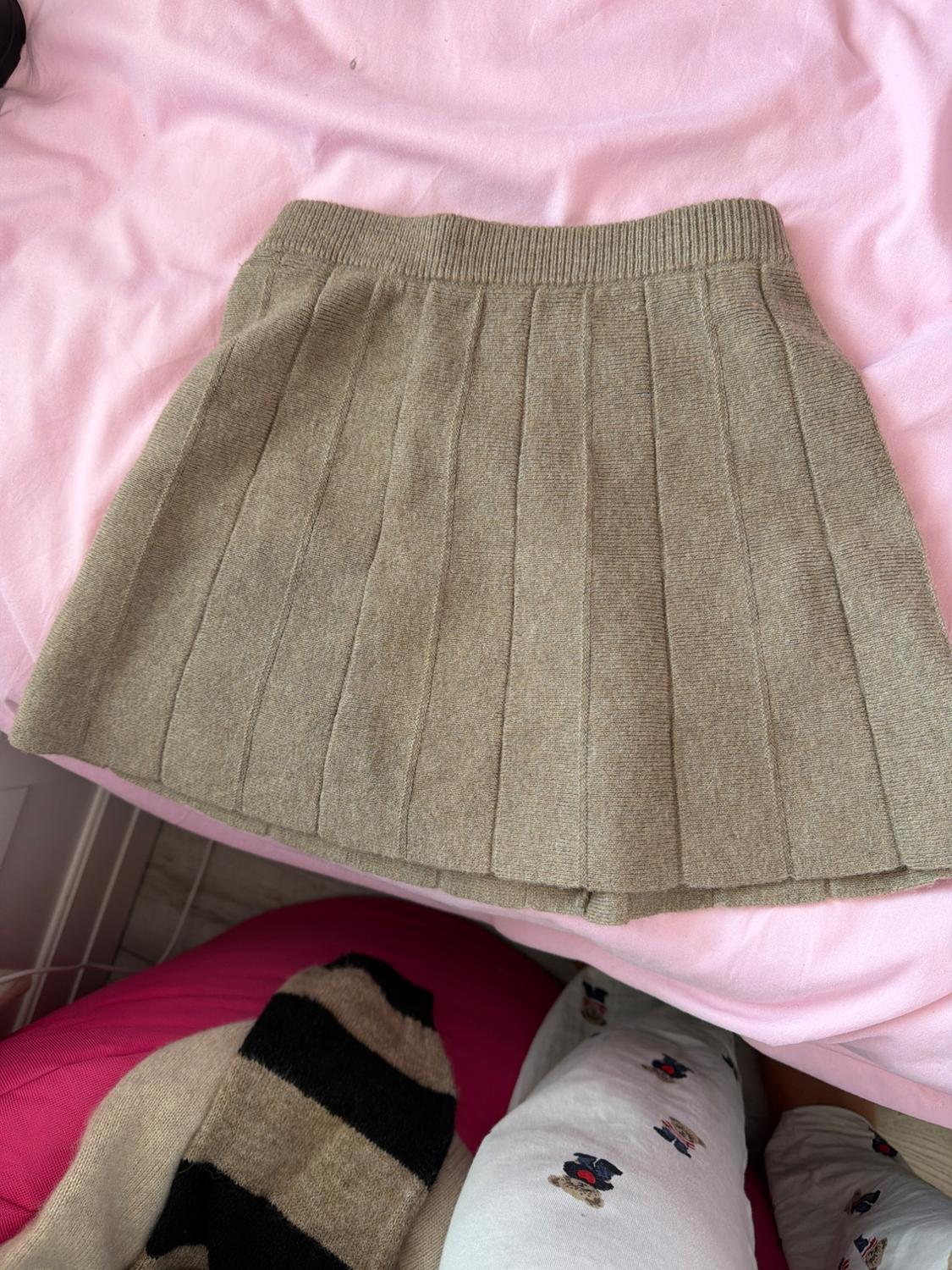 Beige knitted pleated mini skirt over a pink fabric. A striped garment and another with a bear print peek out from the bottom.
