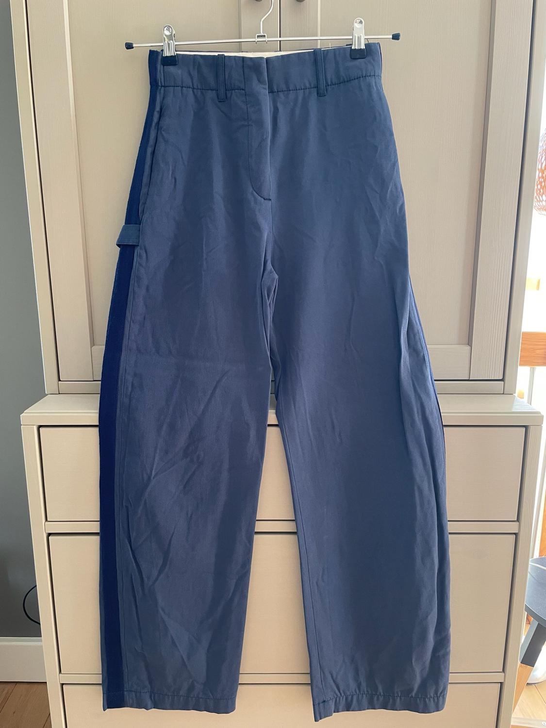 Blue trousers with a dark side stripe hanging on a clothes hanger in front of a chest of drawers.