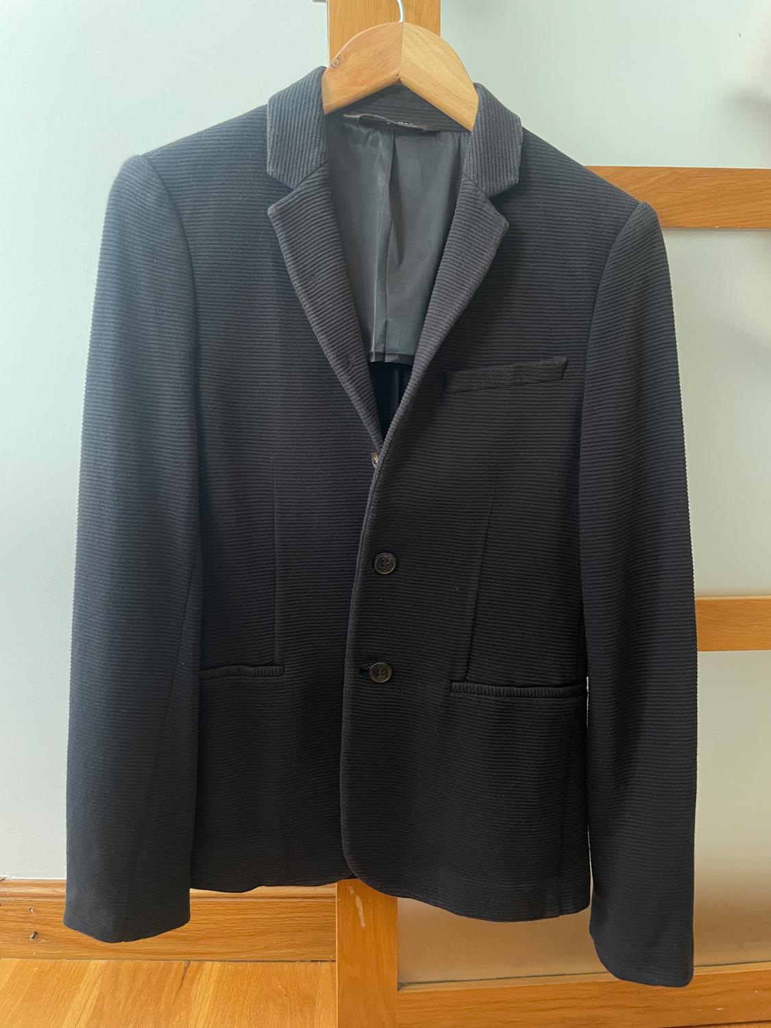 Black corduroy blazer hanging on a hanger over a wooden structure, against a light wall and wooden floor.