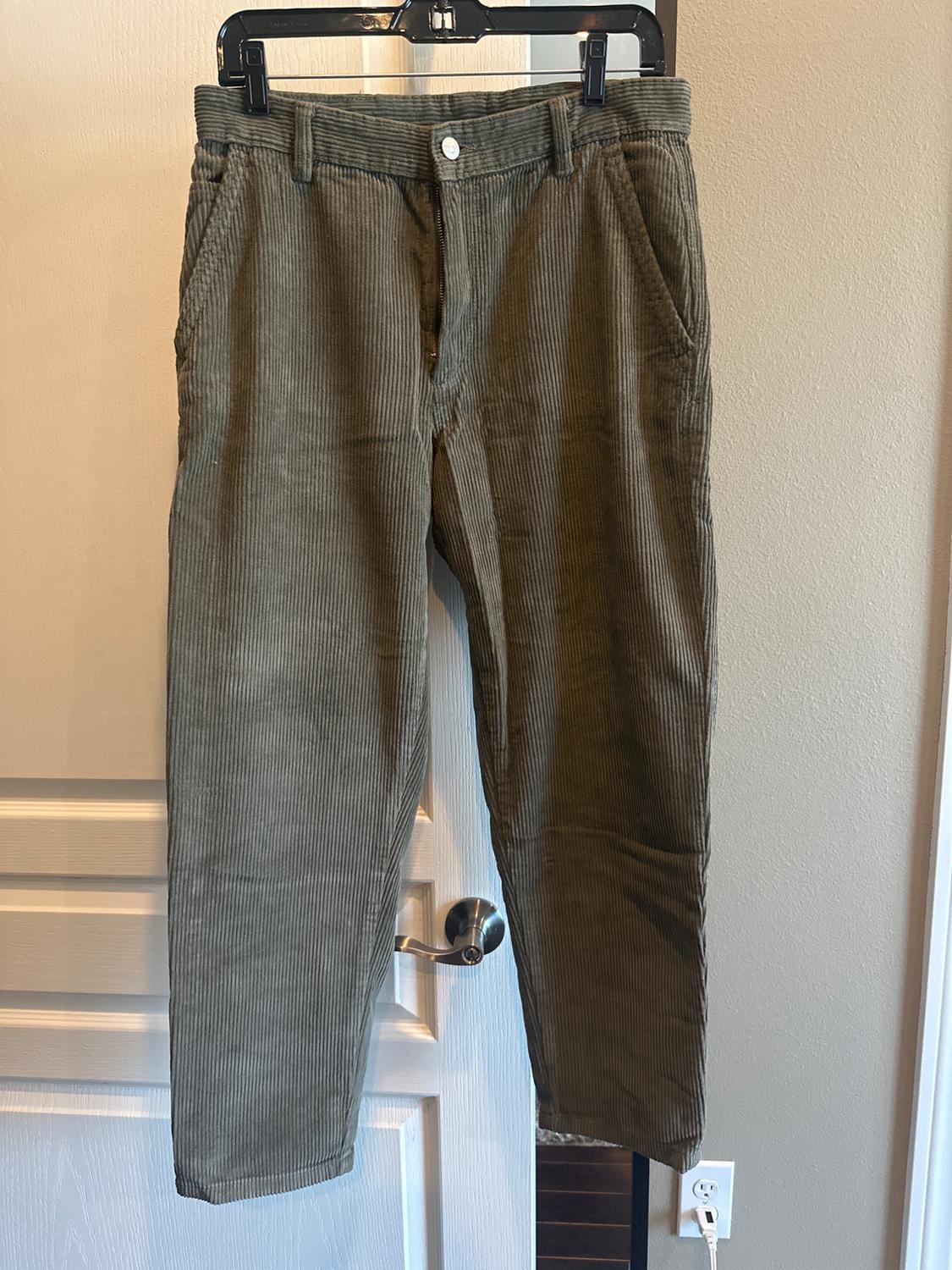 Olive-green corduroy trousers hanging on a black hanger in front of a white door and beige wall. Casual style.