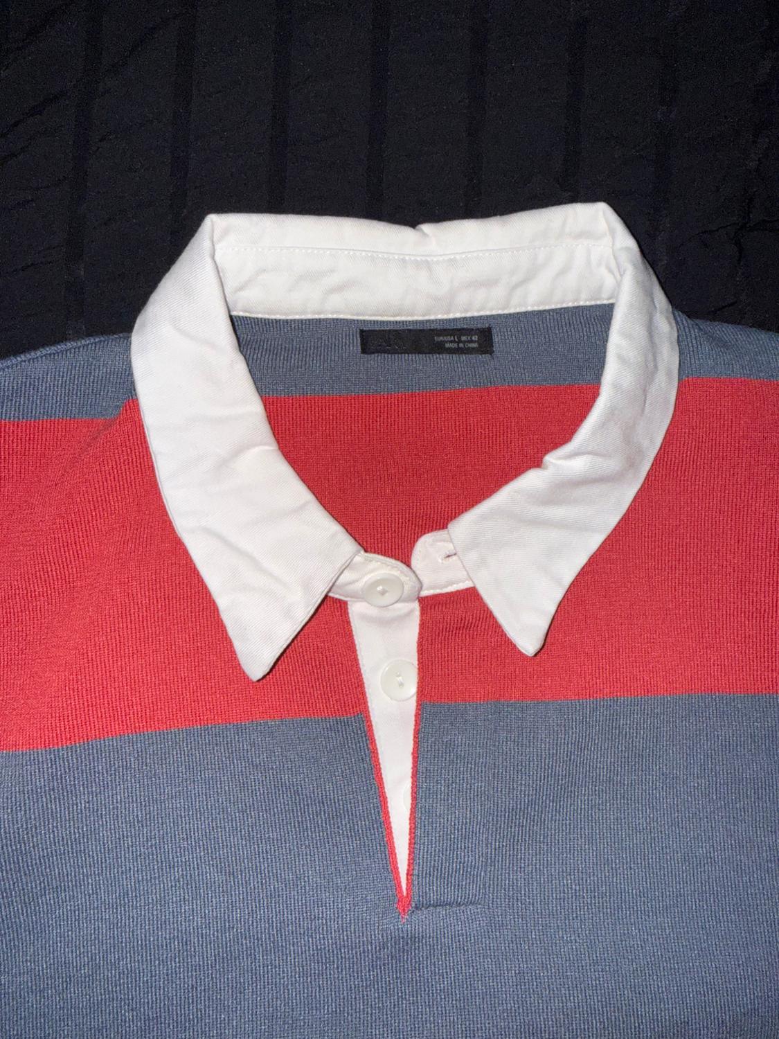 Close-up of a red and gray striped polo shirt with a white collar on black fabric. Text: ZARA EUR/USA L MEX 42 MADE IN CHINA.