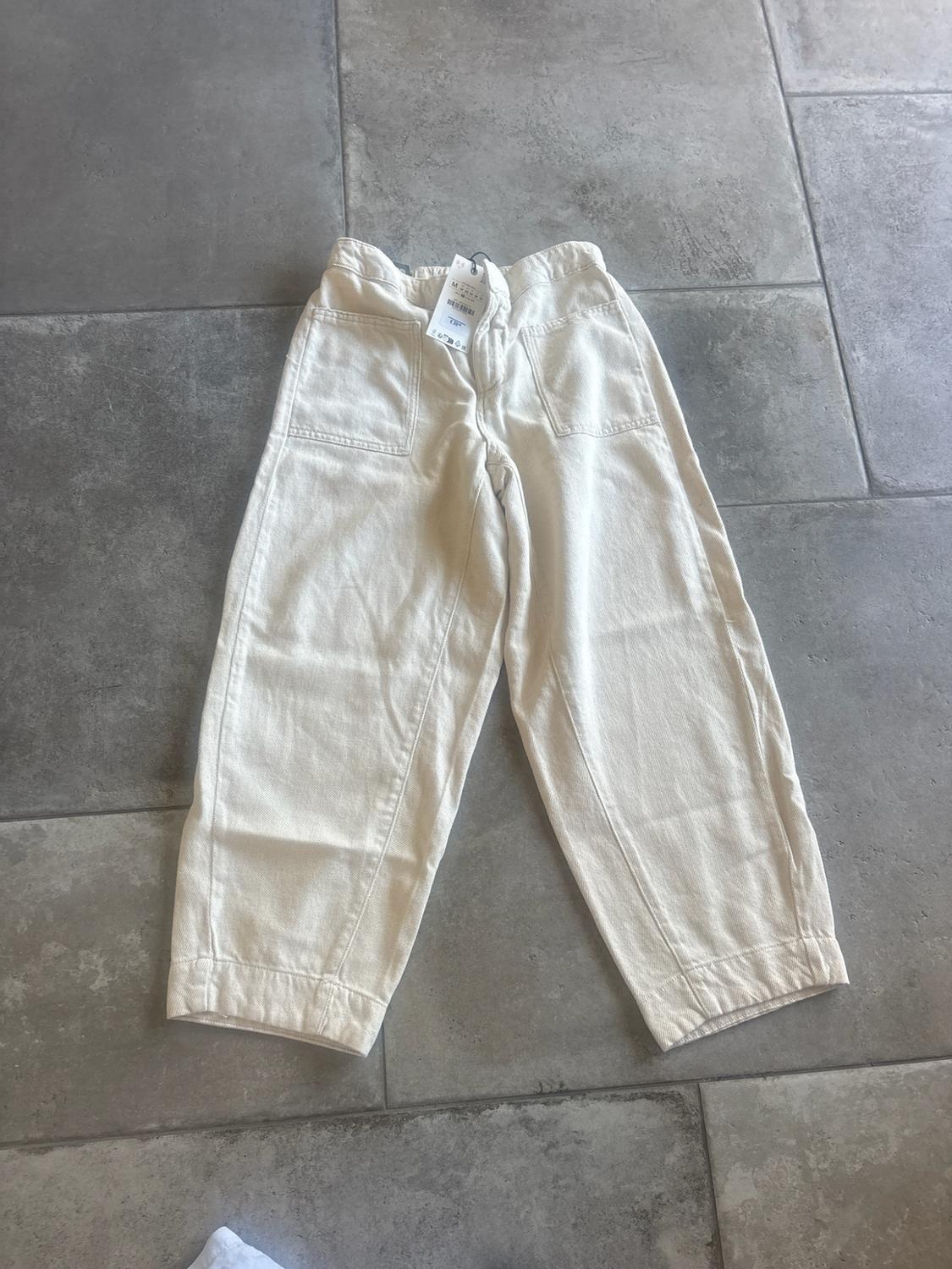 Cream-coloured wide-leg trousers with patch pockets, set against a grey tiled floor. Casual, relaxed style with labels on the waistband.