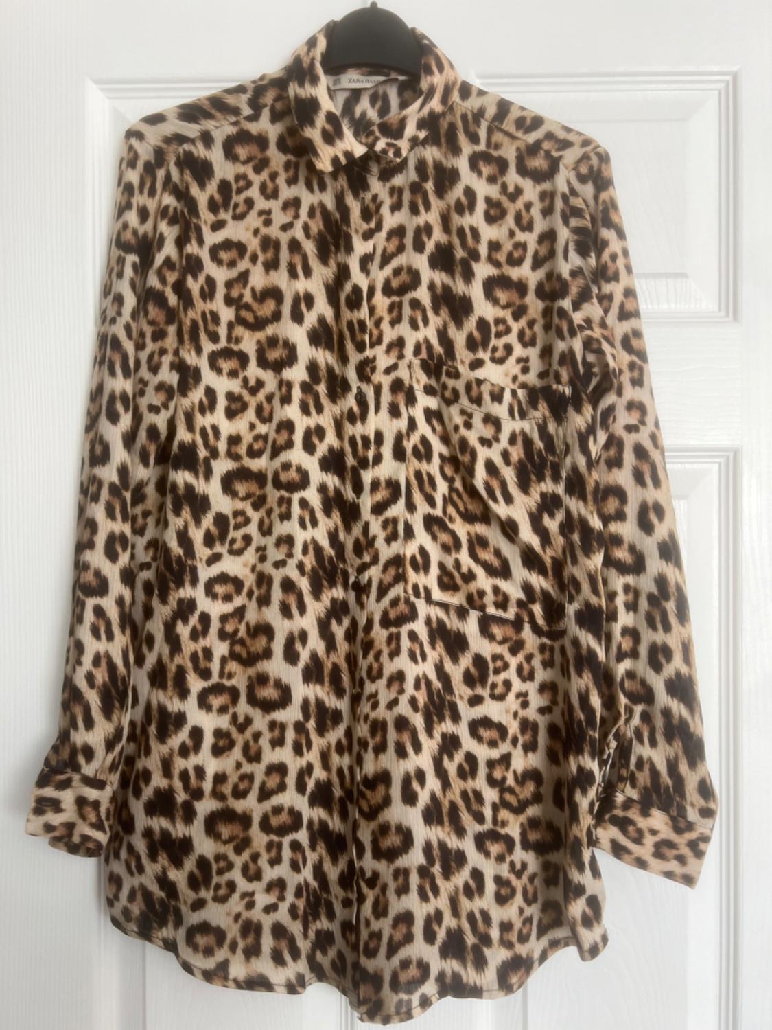 Long-sleeved shirt with a leopard print in shades of brown and beige, hanging on a black hanger in front of a white door.