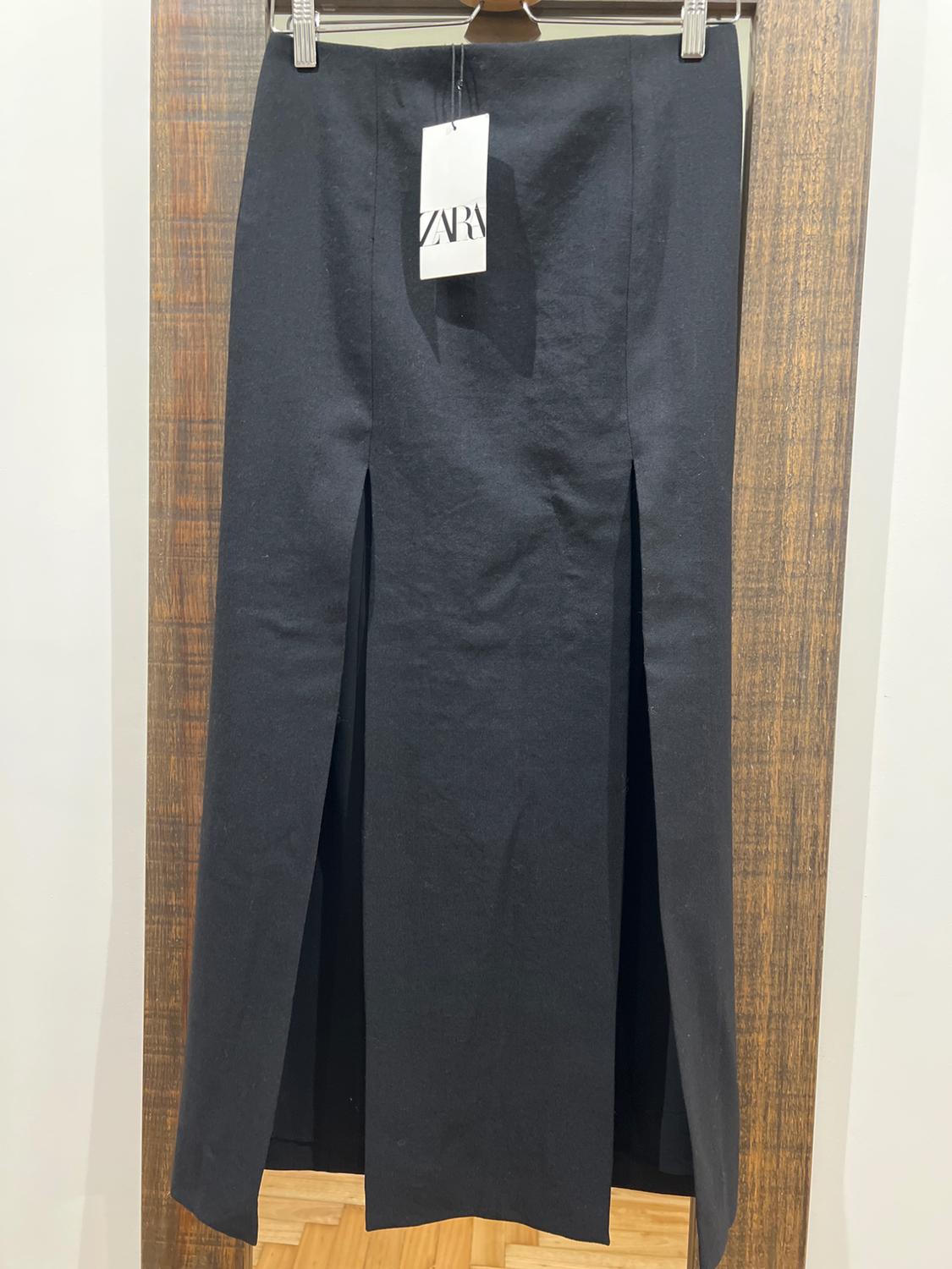 Black skirt with two front slits, hanging on a wooden hanger. Visible label with the text "ZARA". White wall background.