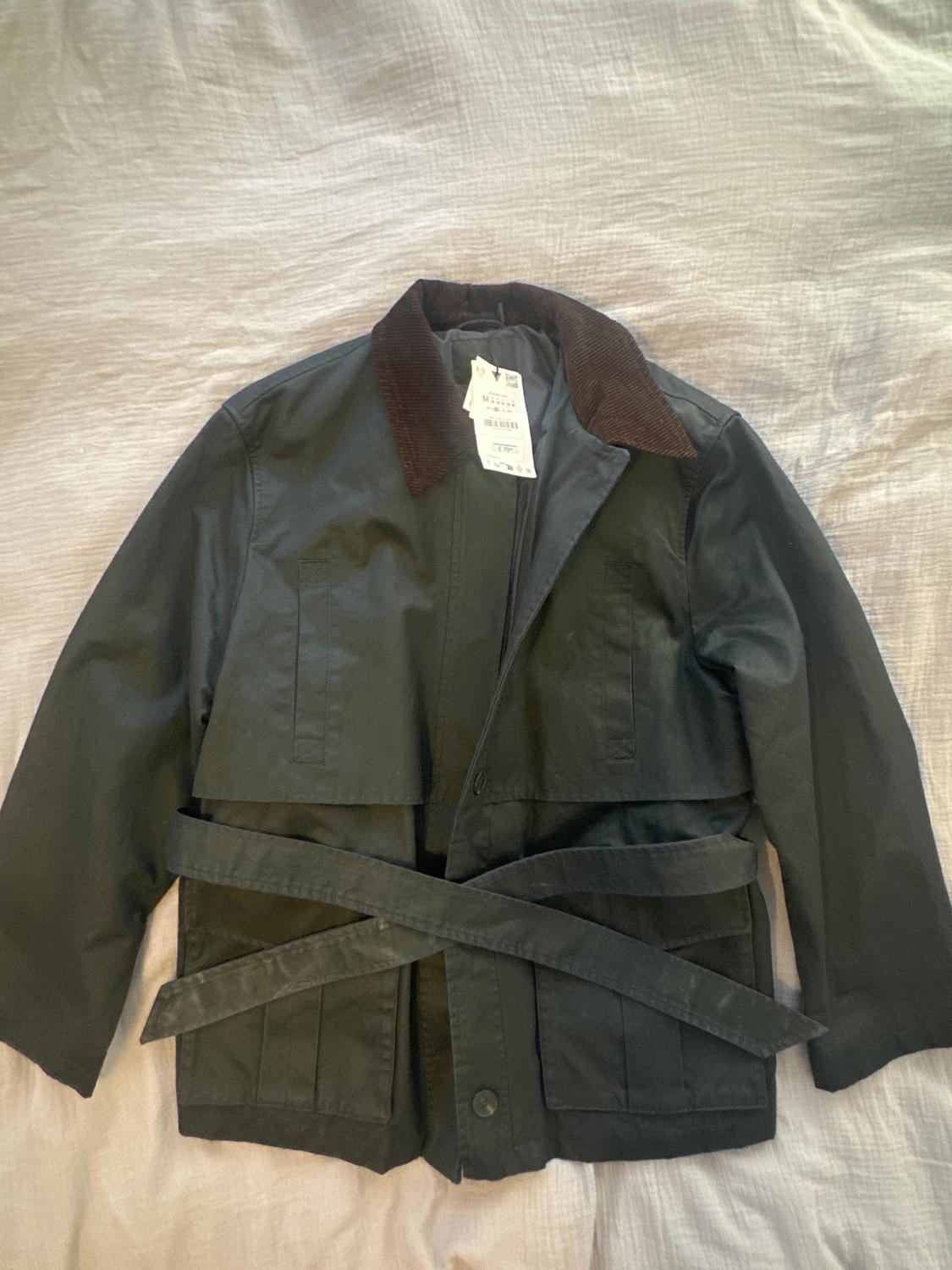 Black jacket with belt and brown collar on a light surface. Tags hanging from the collar.