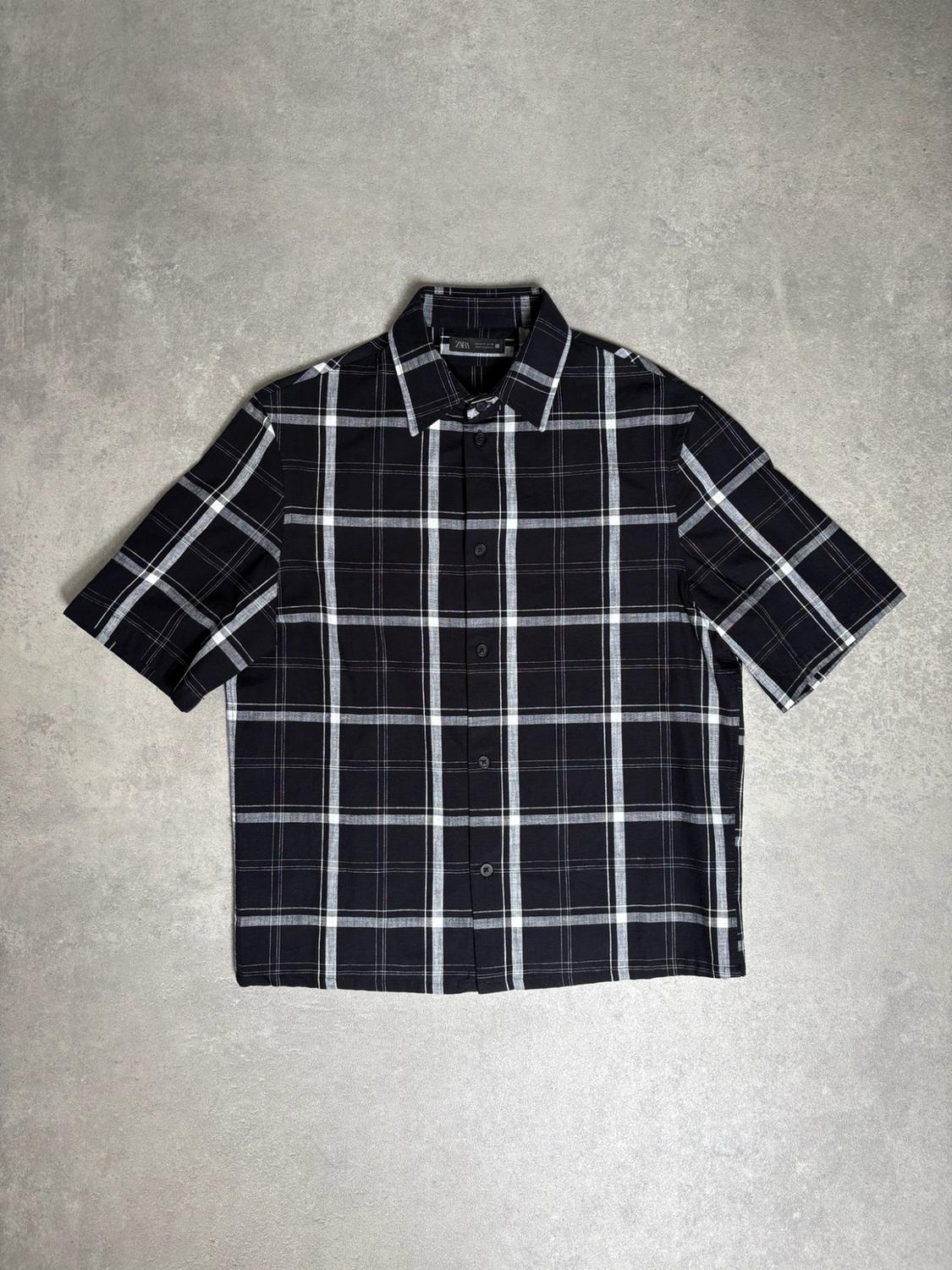 Black short-sleeved shirt with a white check pattern, buttoned, laid out on a textured grey background. Label with the text ZARA.