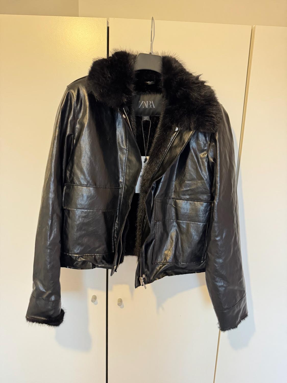 Black jacket with a fur collar and front pockets, hanging in a white wardrobe. Inside label with the text ZARA.