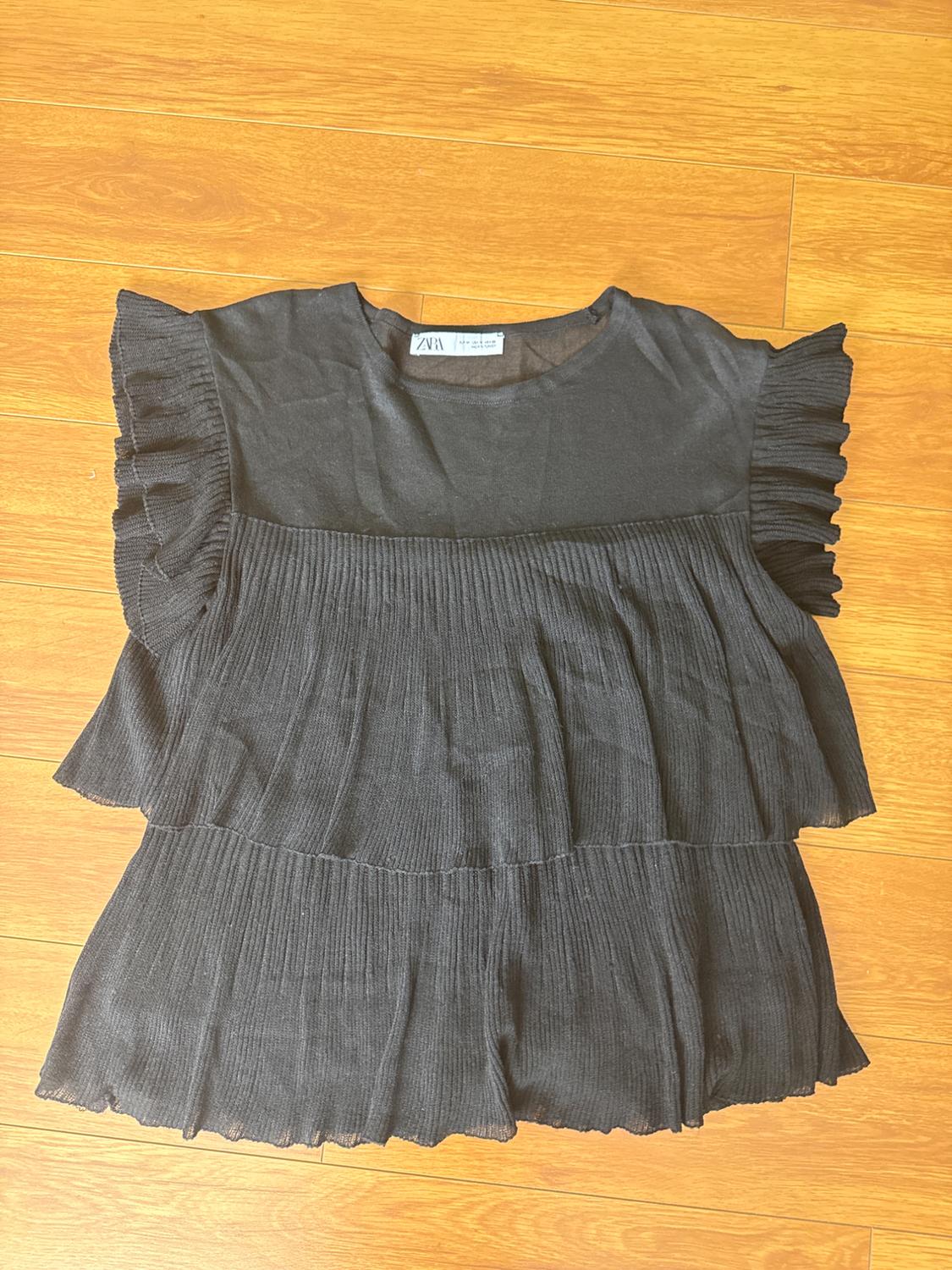 Black blouse with ruffled sleeves and a pleated design, on a light wooden floor. Visible label on the collar.