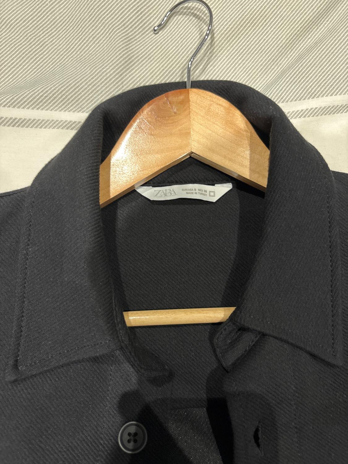 Black shirt hanging on a wooden hanger, with a visible label that says "ZARA" and "MADE IN TURKEY", against a striped fabric background.