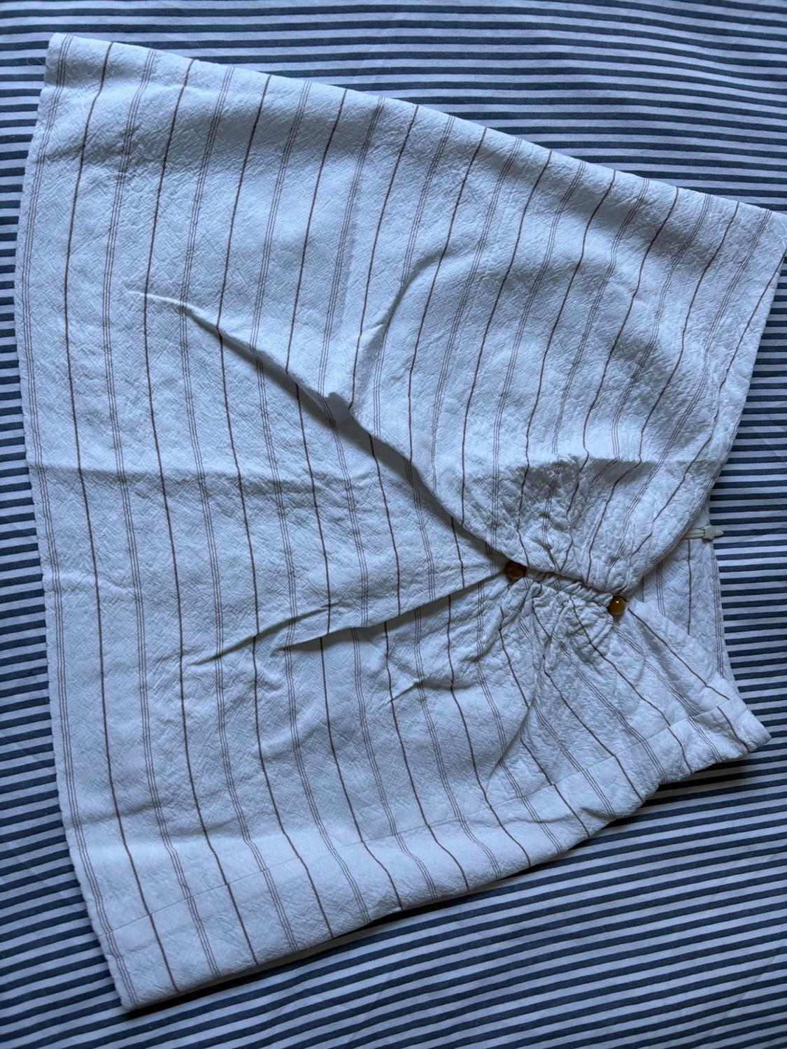 White skirt with brown vertical stripes, featuring gathering and button detail, set against a fabric background with horizontal stripes.