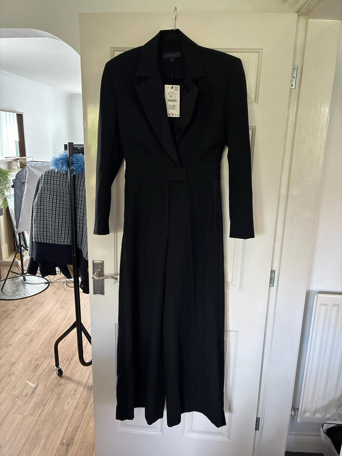 Black long-sleeved jumpsuit with lapel collar hanging on a white door. In the background, a room with a coat rack and wooden floor.