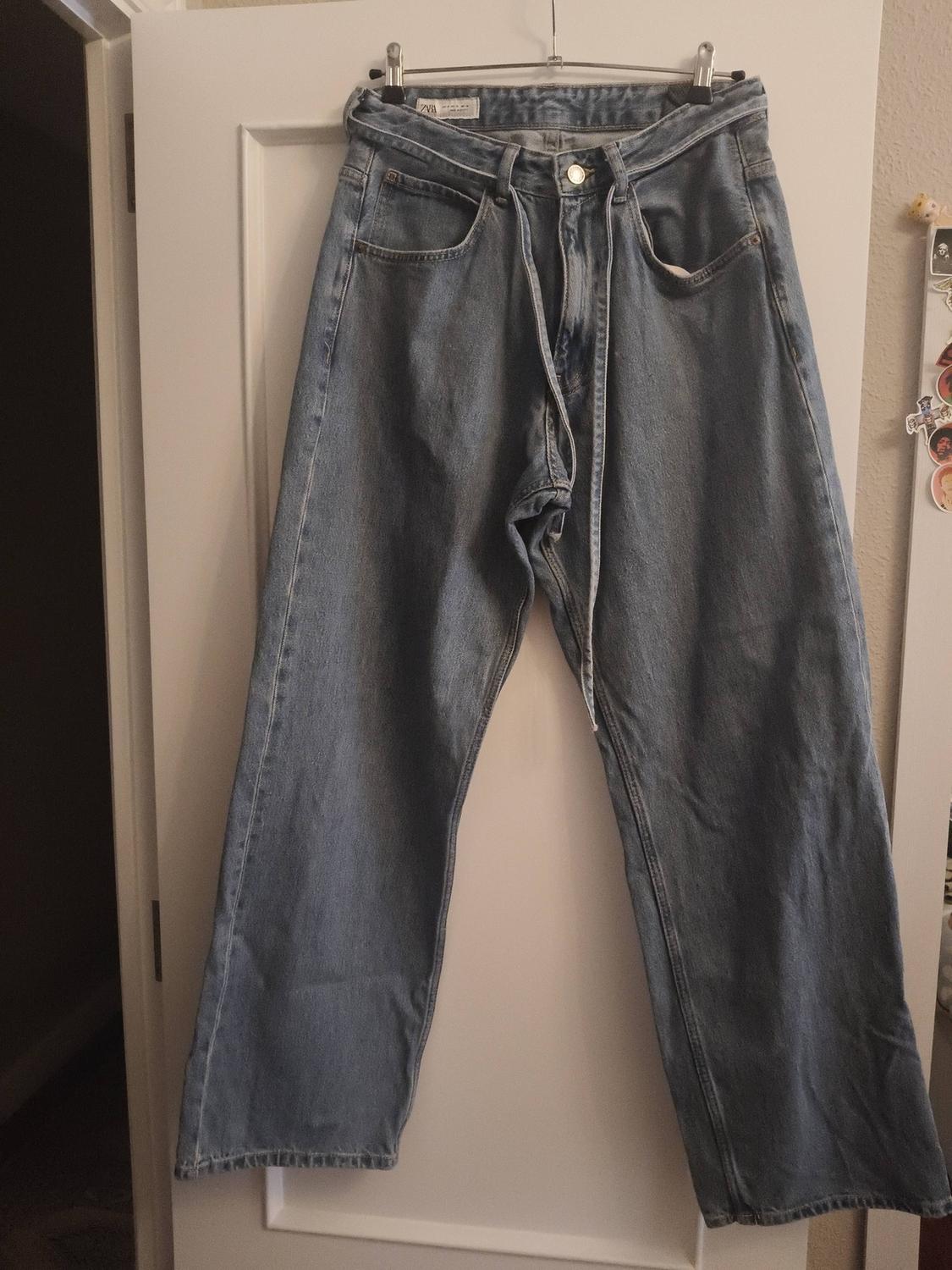Wide-leg blue jeans hanging on a white door. The inner label is visible. Indoor setting with soft light.
