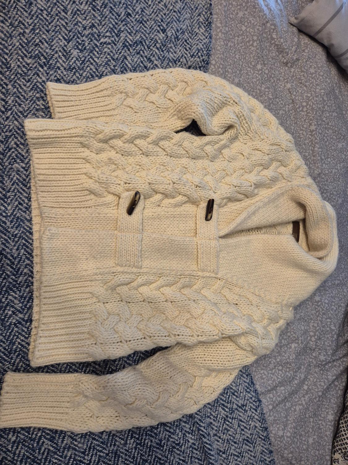 Cream-coloured cable-knit jumper with a shawl collar and wooden buttons, on a bed with patterned blankets in shades of blue and grey.