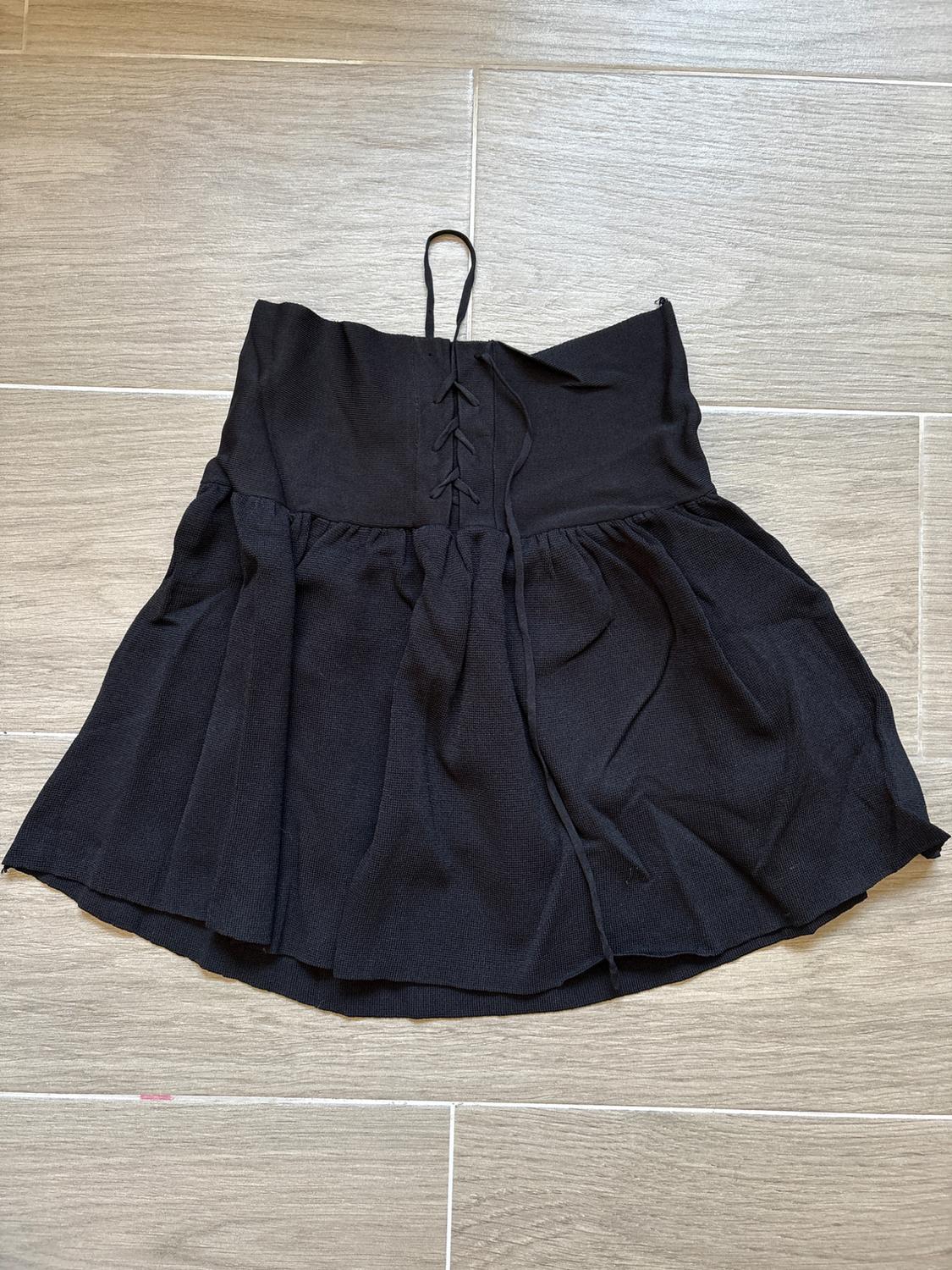 Black mini skirt with pleated design and adjustable drawstrings at the top, placed on a light grey tiled floor.