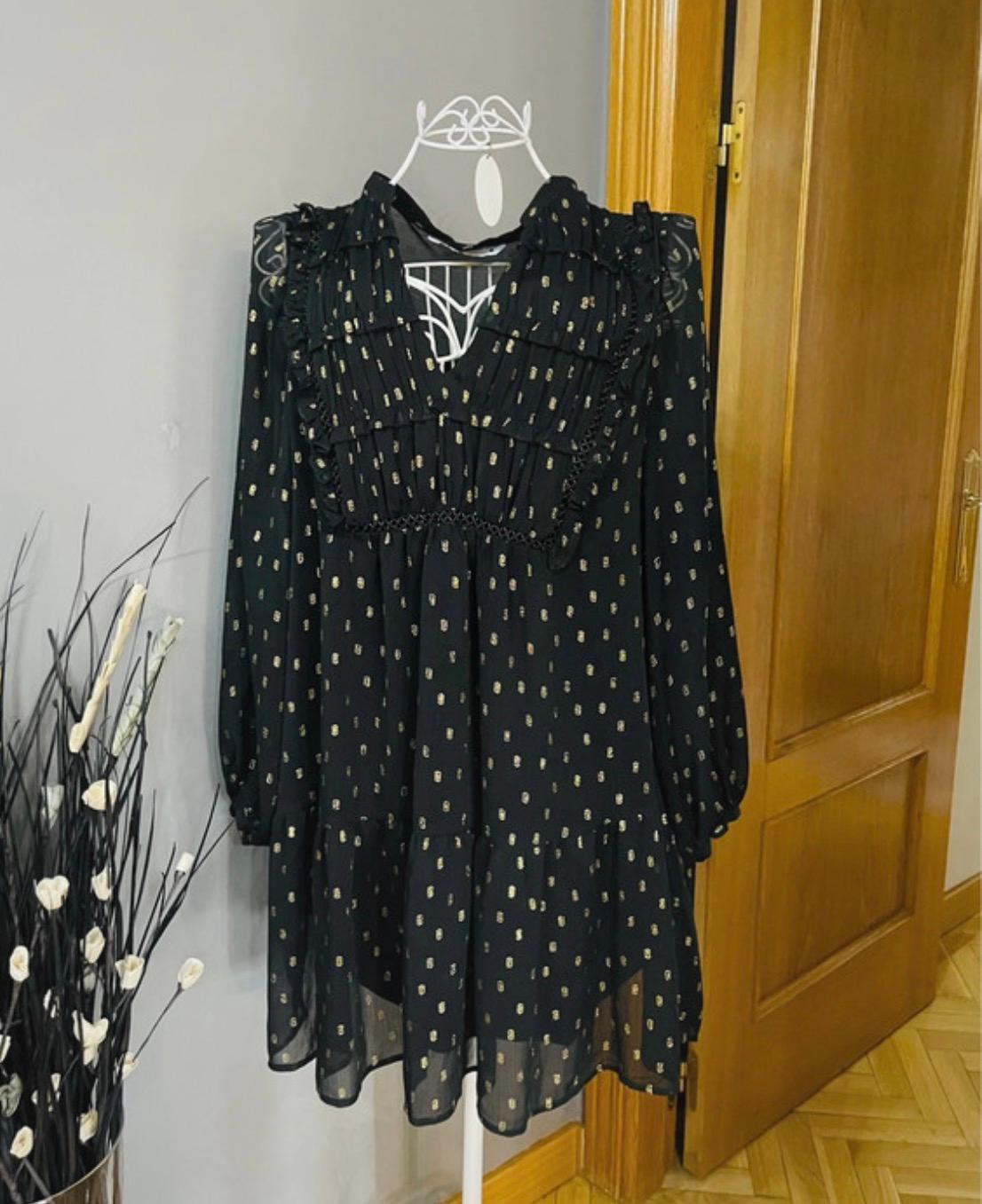 Black dress with gold polka dot print, long sleeves and ruffles, hanging on a white clothes rail in a room with a wooden door.