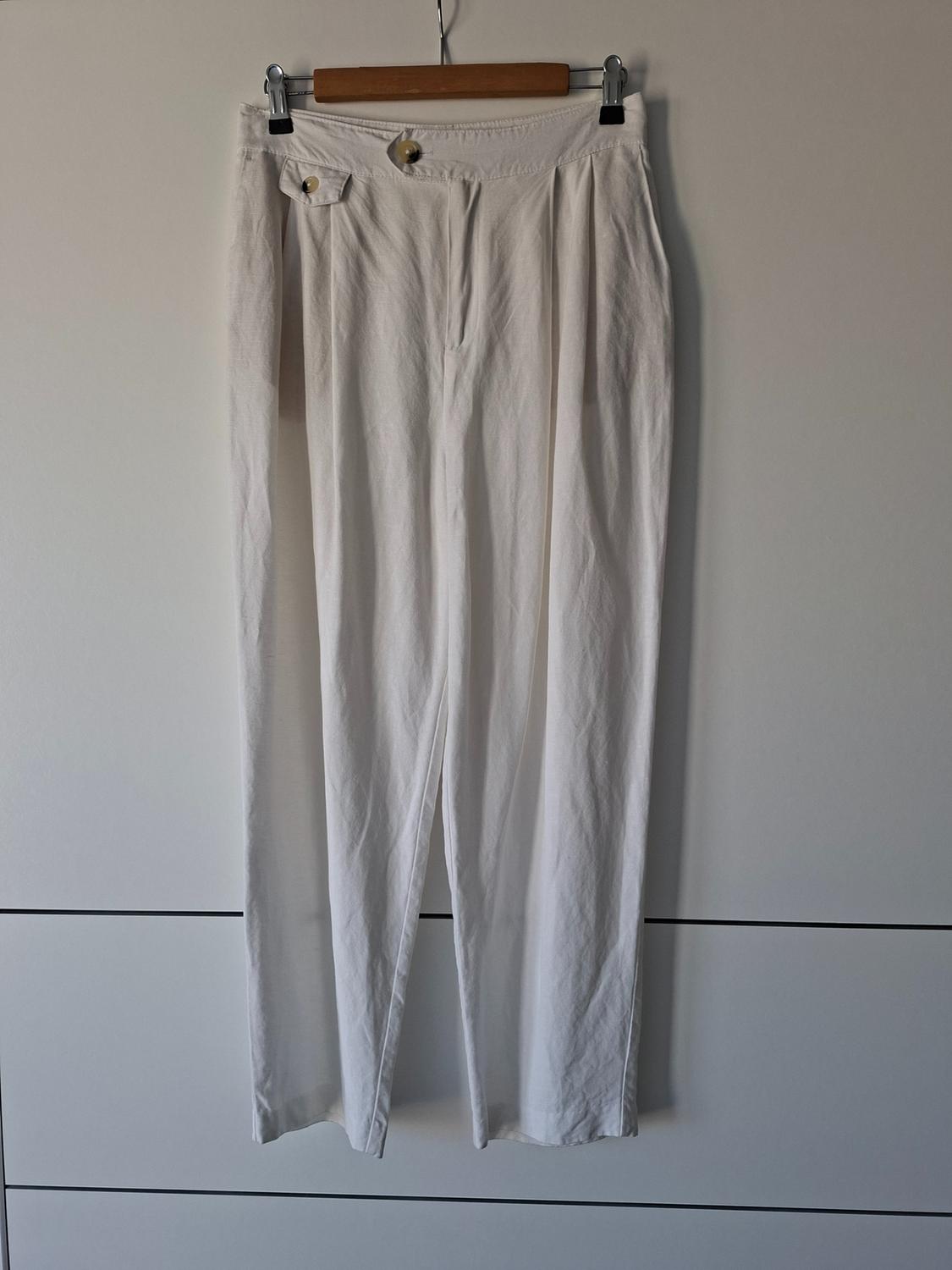 White trousers with pleats and visible buttons, hanging from a wooden hanger against a white background with black horizontal lines.