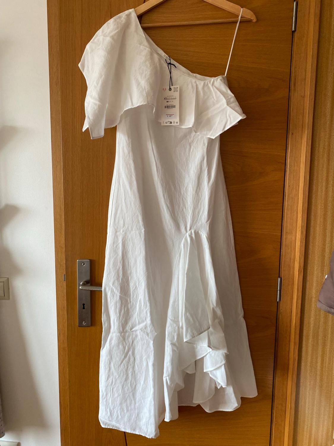 Asymmetric white dress with ruffles hanging on a wooden door. Visible label with the text XS.