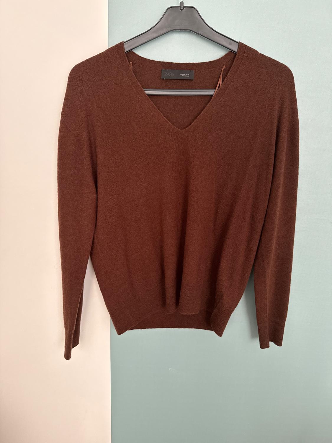 Brown long-sleeved V-neck jumper on a black hanger. Background split into cream and light blue. ZARA text on the label.