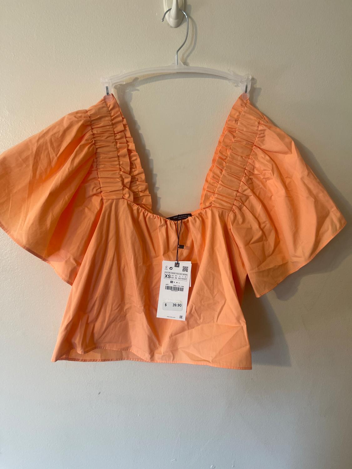 Orange blouse with puff sleeves and a V-neck, hanging on a hanger. Visible tag with price of $39.90 and size XS.