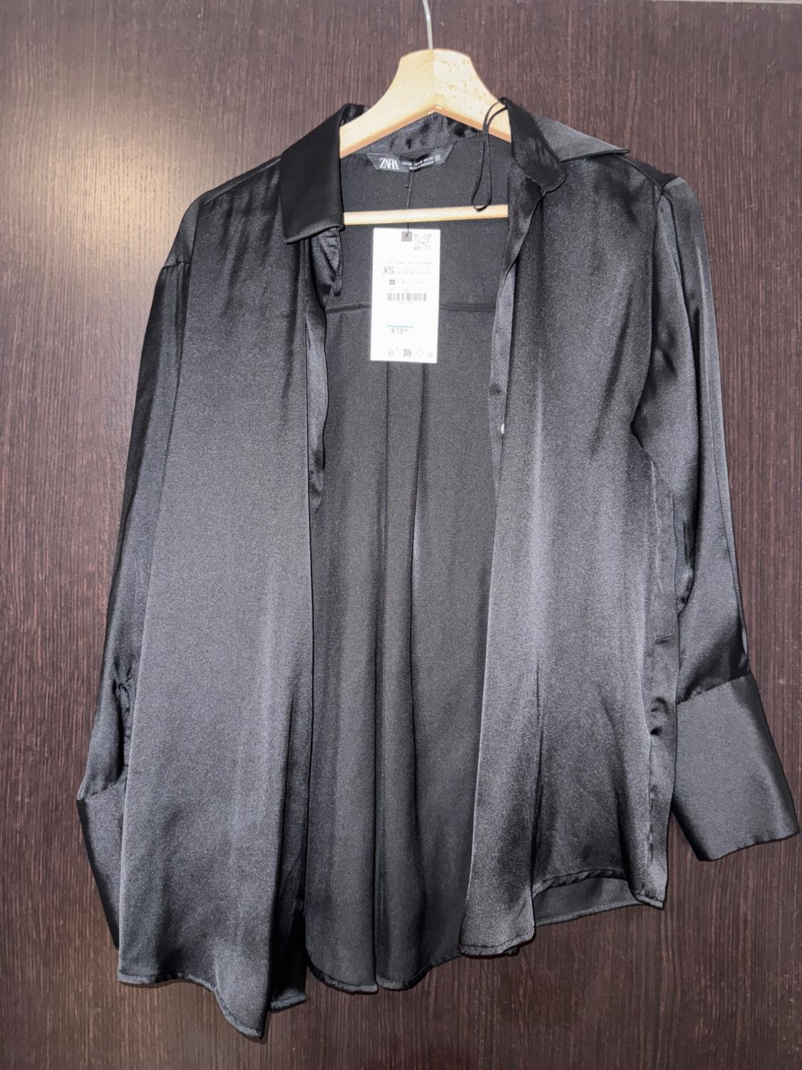Black satin shirt hanging on a wooden hanger, with a visible label in the centre, against a dark wooden background.