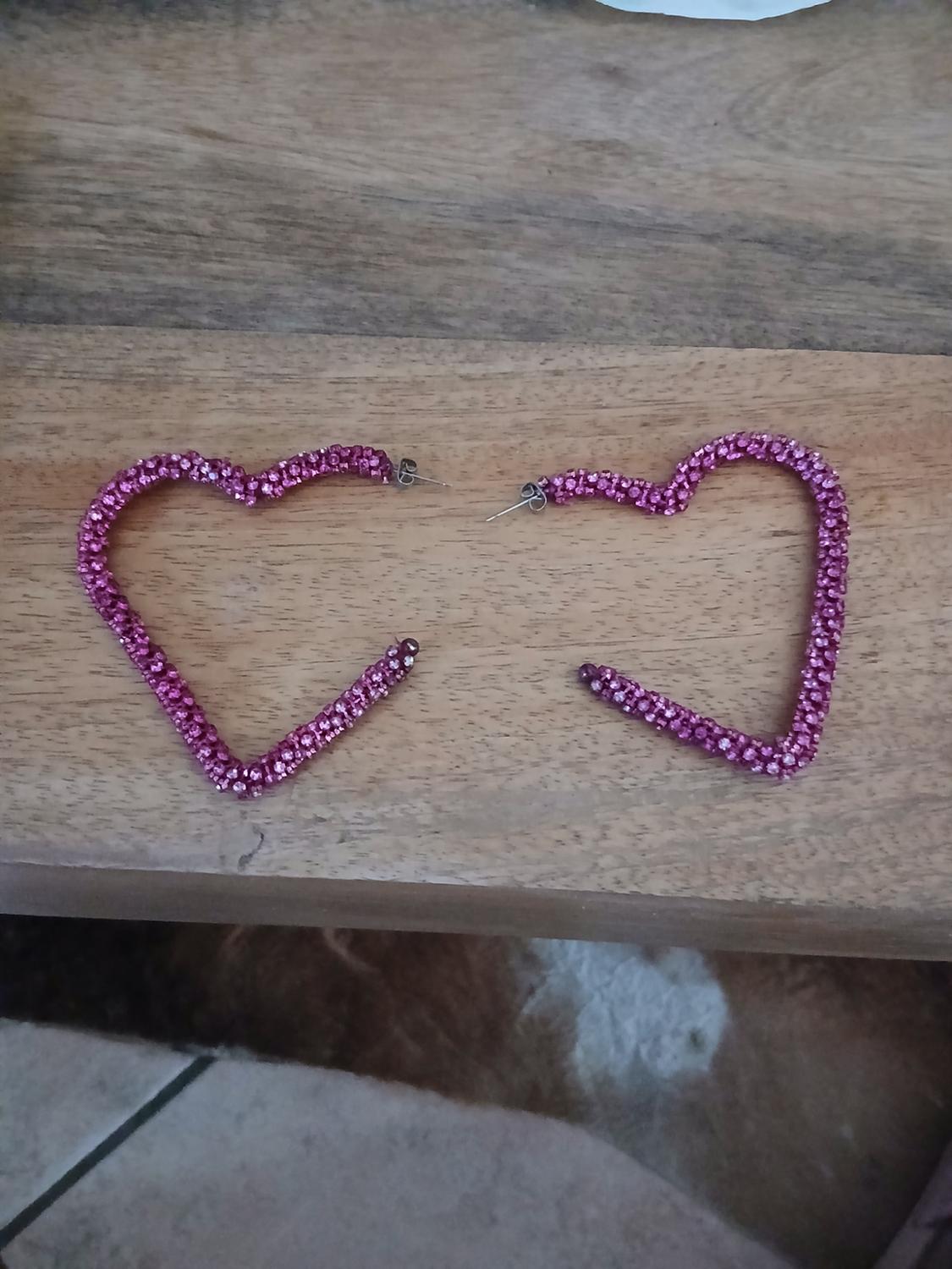 Two open heart-shaped earrings, pink with sparkling rhinestones, resting on a wooden surface.