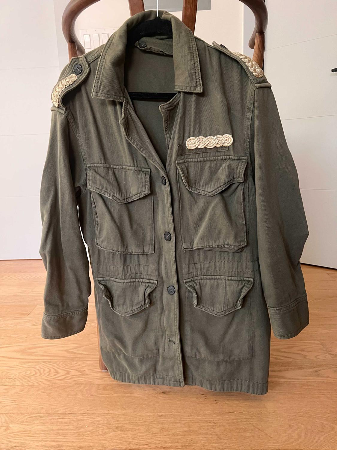 Olive green military jacket hanging on a wooden chair. Its four pockets and braided gold details on the shoulders and chest stand out.