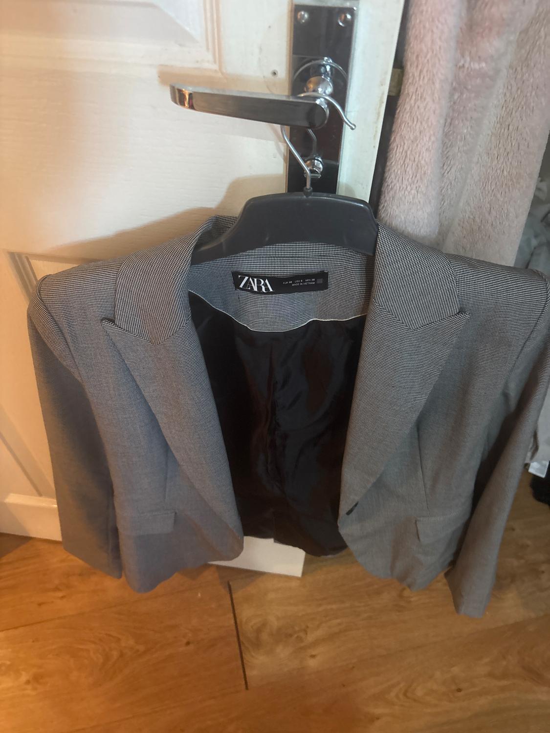 Grey blazer with black lining and ZARA label, on a hanger on the knob of a white door. Wooden floor, casual setting.