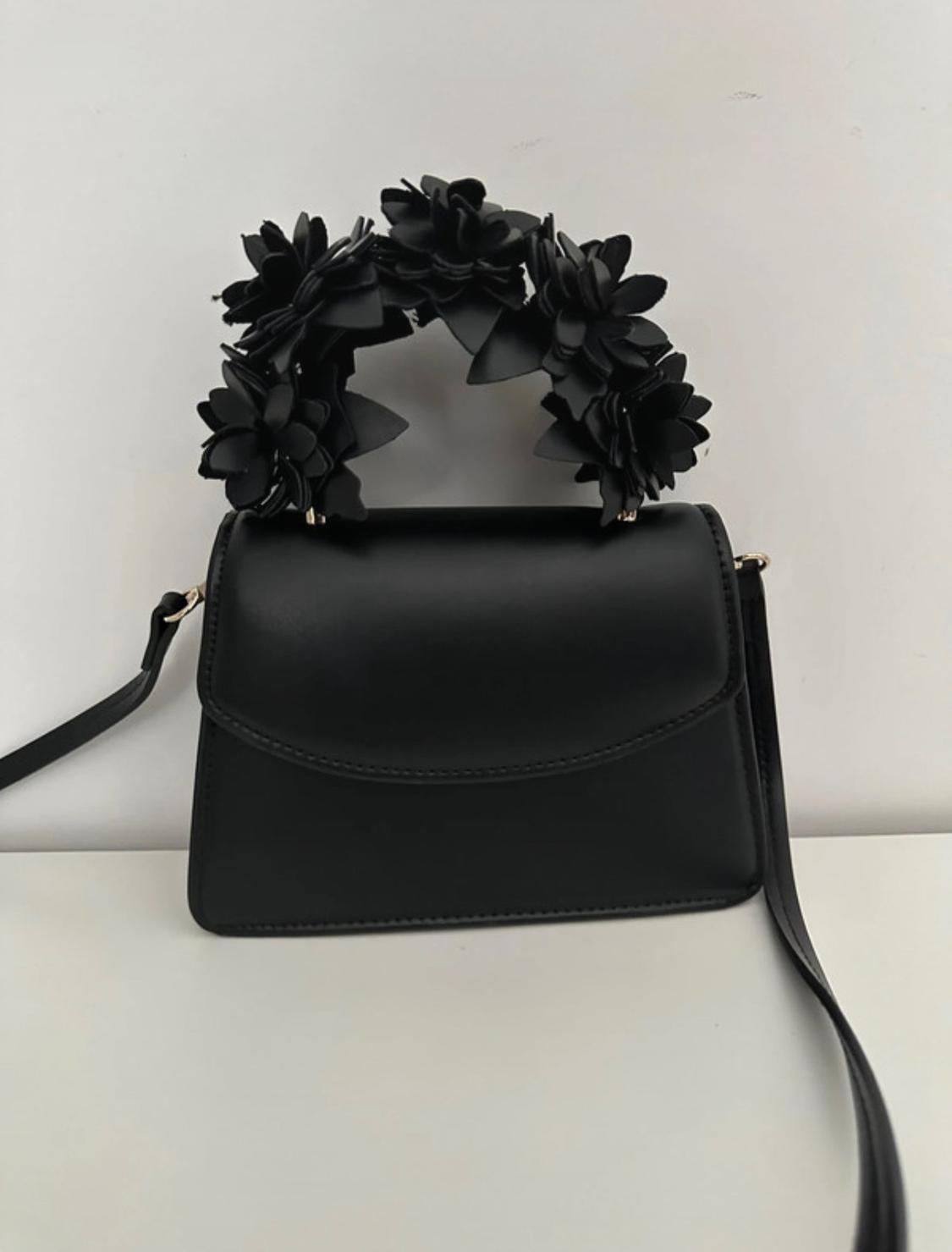 Black flap bag with a long strap. Featuring a distinctive handle covered in black flowers. Minimalist composition on a white background.