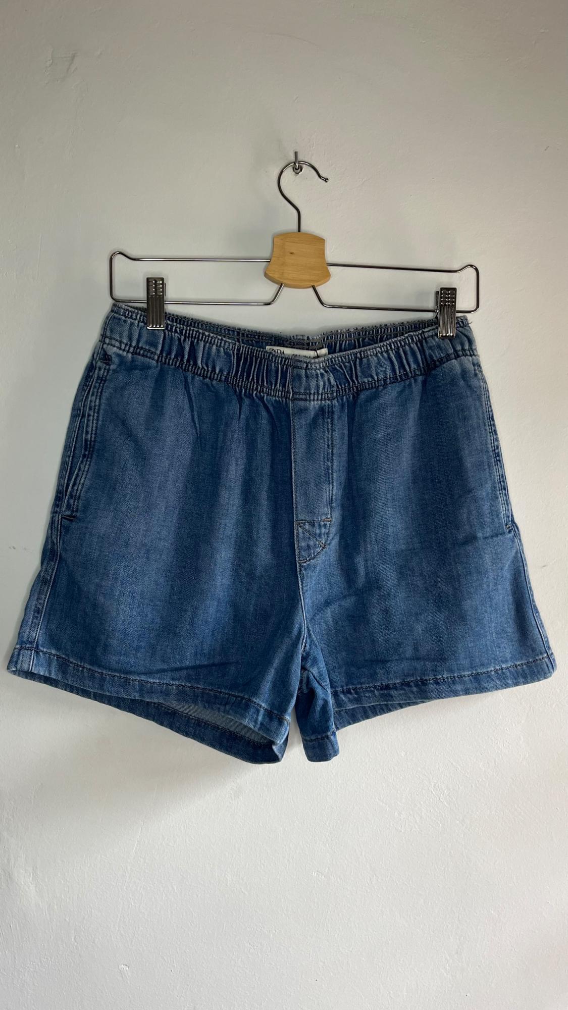 Blue denim shorts hanging on a metal hanger against a white background. The design is simple with an elasticated waist.