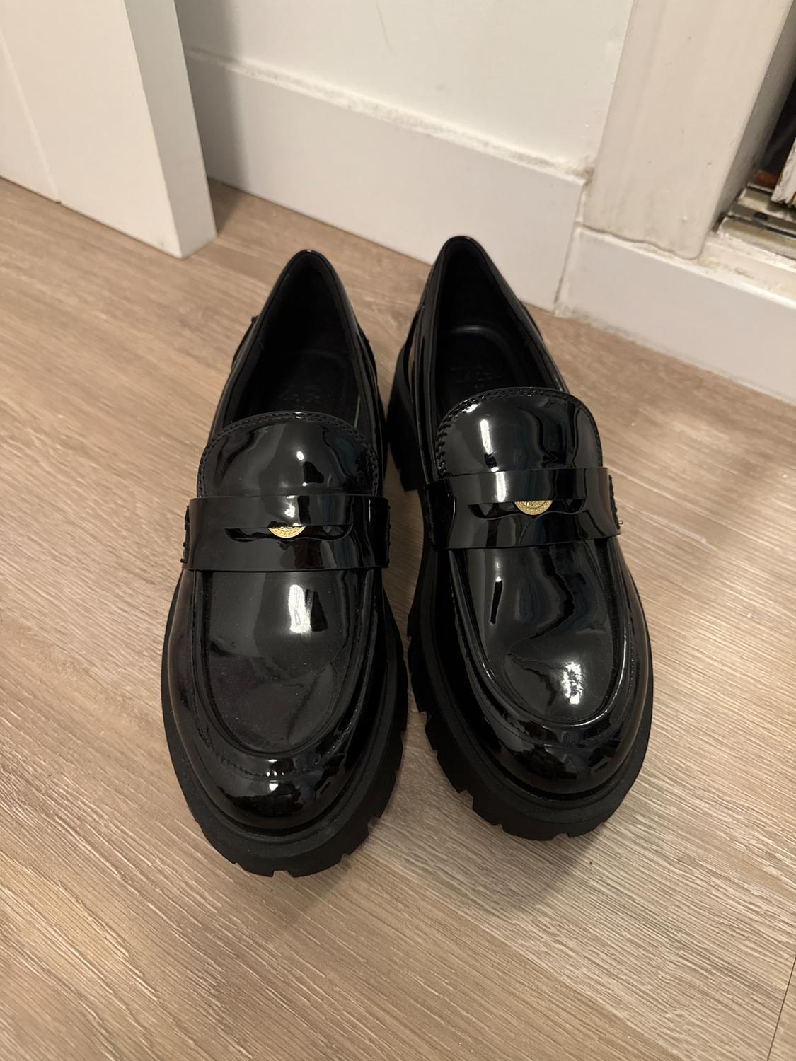 Black patent leather shoes with a thick sole on a light wooden floor, viewed from above in an indoor setting.